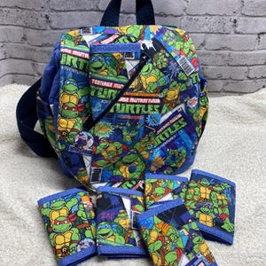 May include: A blue backpack and matching wallets feature a colorful Teenage Mutant Ninja Turtles comic book design. The backpack has a top handle and adjustable straps. The wallets are rectangular with a blue border and the same comic book print.