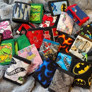 May include: A collection of 18 small quilted wallets with various colorful patterns and designs. The wallets feature a variety of themes, including cartoon characters, sports teams, and abstract designs. The wallets are made of fabric and have a black trim.