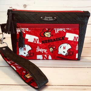 May include: Black and red zippered pouch with a black woven strap and tassel. The pouch features a red and white fabric with the words "Nebraska" and "Huskers" and a cartoon mascot. The pouch has a black label with the text "Country Chic Sews".