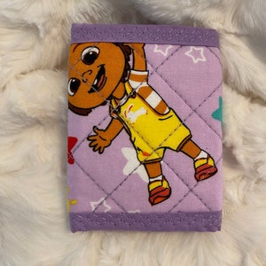 May include: A purple quilted wallet with a cartoon character on the front. The character is a brown-skinned girl with yellow clothing and a white star pattern on the background.