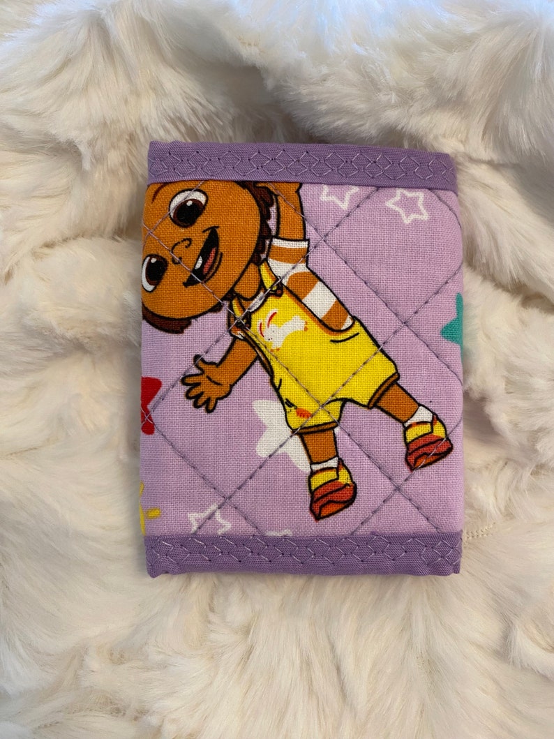 May include: A purple quilted wallet with a cartoon character on the front. The character is a brown-skinned girl with yellow clothing and a white star pattern on the background.