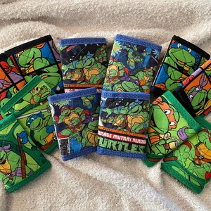 May include: Nine Teenage Mutant Ninja Turtles themed fabric wallets with green, black, and blue accents. The wallets feature different images of the turtles and the text "Teenage Mutant Ninja Turtles".