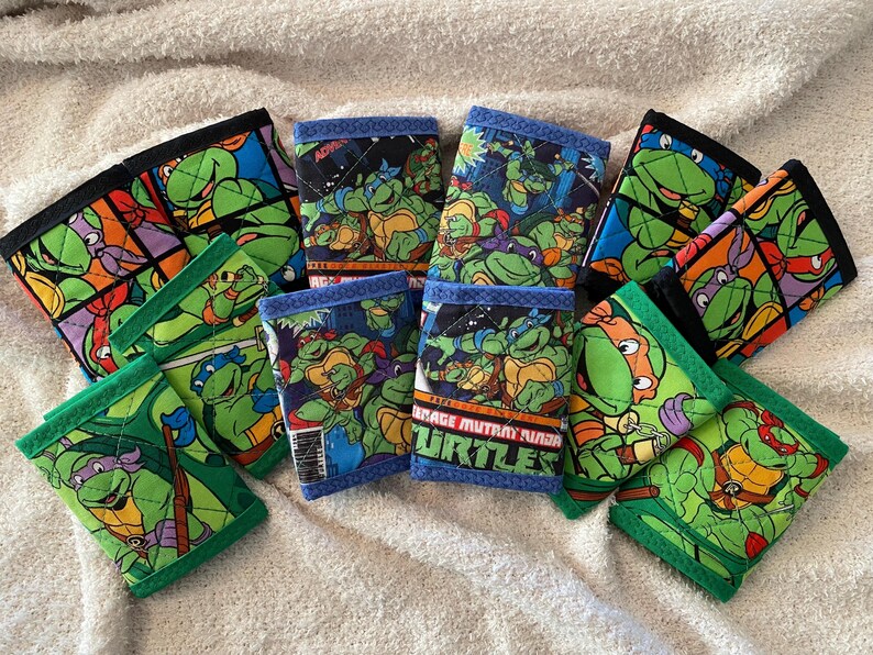 May include: Nine Teenage Mutant Ninja Turtles themed fabric wallets with green, black, and blue accents. The wallets feature different images of the turtles and the text "Teenage Mutant Ninja Turtles".
