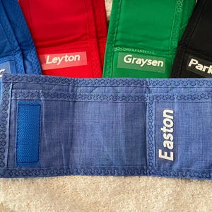 May include: Blue, red, green, and black fabric wallets with white personalized names: Zedric, Leyton, Graysen, and Parker. The blue wallet has a blue strap and the name Easton.