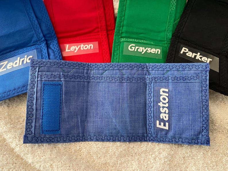 May include: Blue, red, green, and black fabric wallets with white personalized names: Zedric, Leyton, Graysen, and Parker. The blue wallet has a blue strap and the name Easton.