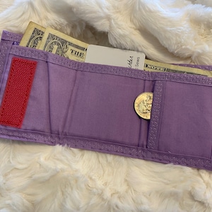 May include: A purple fabric wallet with a red velcro closure, a coin pocket, and a billfold. The wallet contains cash and a credit card.
