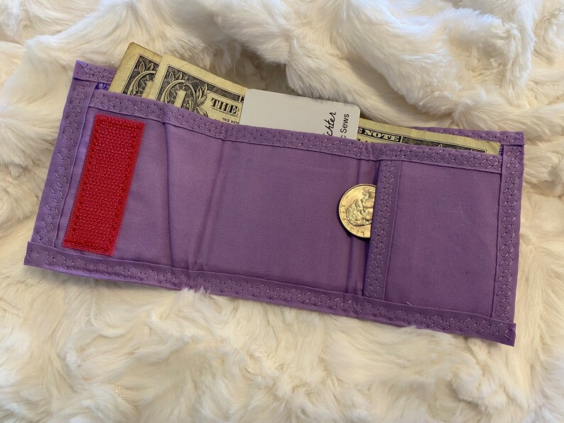 May include: A purple fabric wallet with a red velcro closure, a coin pocket, and a billfold. The wallet contains cash and a credit card.