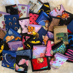 May include: A collection of colorful quilted fabric wallets with various cartoon and character designs, including a tractor, a horse, a butterfly, a cat, a princess, a superhero, and a truck.