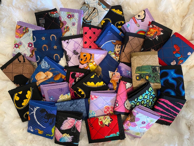 May include: A collection of colorful quilted fabric wallets with various cartoon and character designs, including a tractor, a horse, a butterfly, a cat, a princess, a superhero, and a truck.