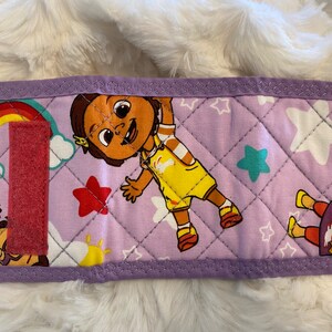 May include: Purple quilted fabric pouch with a pink velcro closure. The pouch features a colorful design with a cartoon character, stars, and a rainbow.