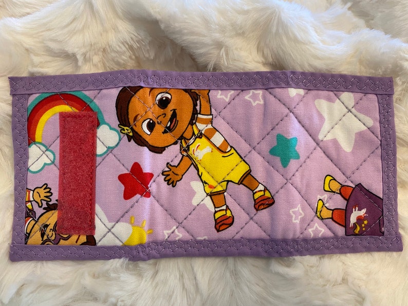 May include: Purple quilted fabric pouch with a pink velcro closure. The pouch features a colorful design with a cartoon character, stars, and a rainbow.