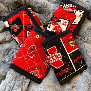 May include: Four red and black quilted fabric wallets with different Nebraska Huskers designs. The wallets feature a variety of logos, including the Husker 'N', the Husker 'I', and the Husker 'H'.