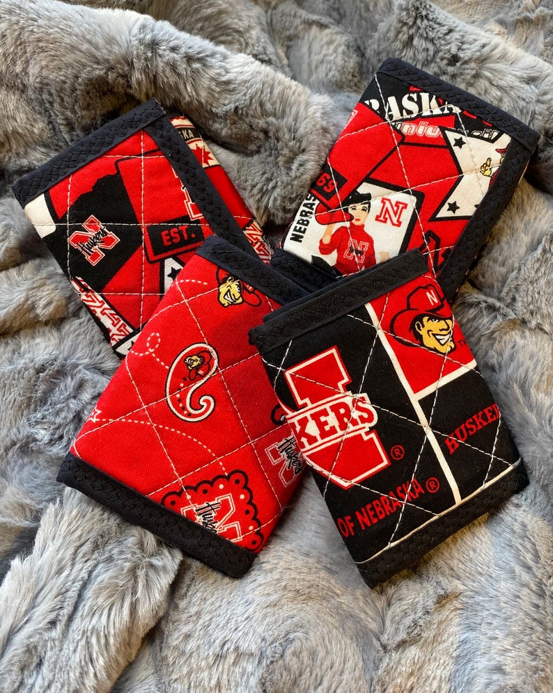May include: Four red and black quilted fabric wallets with different Nebraska Huskers designs. The wallets feature a variety of logos, including the Husker 'N', the Husker 'I', and the Husker 'H'.