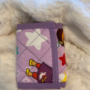 May include: A purple quilted wallet with a cartoon girl and stars design. The wallet has a purple trim and a white star in the center.