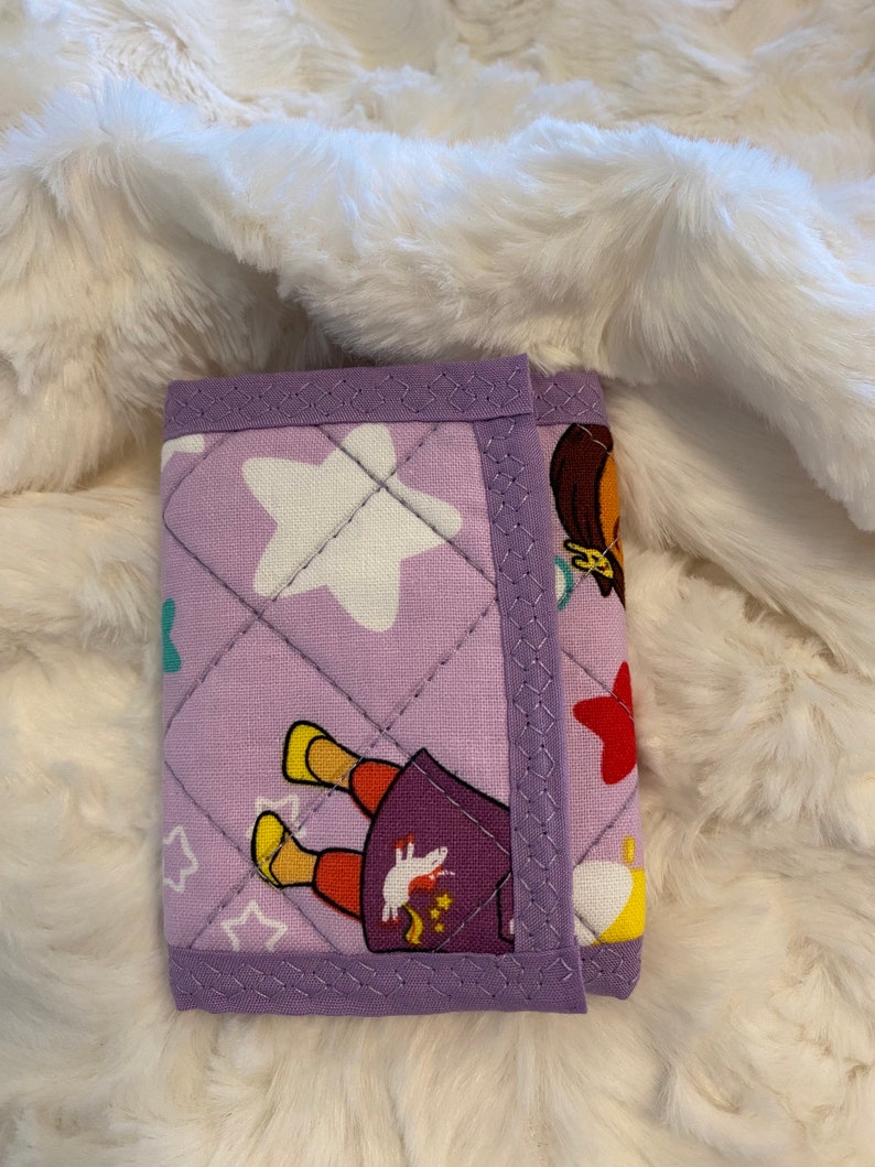 May include: A purple quilted wallet with a cartoon girl and stars design. The wallet has a purple trim and a white star in the center.