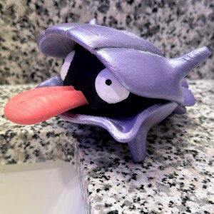May include: A purple and black 3D printed Pokemon figurine of a Cloyster. The figurine has a large, open shell with a black interior and a red tongue sticking out.