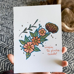 May include: A white card with a floral illustration and the text "limpia tu pinche cuarto!" in Spanish. The flowers are pink, orange, and brown with green leaves. The background is white with a subtle texture.
