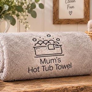 May include: A rolled light grey bath towel featuring an embroidered black graphic of a hot tub with bubbles and the text Mum's Hot Tub Towel. The towel has a soft, textured fabric surface and is positioned horizontally on a wooden surface.