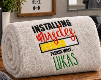 Può includere: Un asciugamano beige arrotolato con testo ricamato che recita INSTALLING muscles PLEASE WAIT... LUKAS. Il design include una grafica a barra di caricamento con segmenti gialli. Il testo è realizzato con filo nero, rosso e verde sul tessuto.