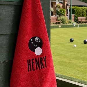 May include: A bright red towel featuring an embroidered design of a black lawn bowl and a white jack ball. Below the graphic, the name HENRY is embroidered in black capital letters. The towel is hanging from a hook on a dark green wooden post.