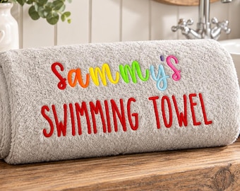 Könnte beinhalten: Ein gerolltes hellgraues Handtuch mit gesticktem Text. Das Wort Sammy's ist in einem mehrfarbigen Regenbogenverlauf gestickt, während die Worte SWIMMING TOWEL in fetten roten Großbuchstaben in der Mitte des Stoffes eingestickt sind.