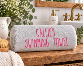 Könnte beinhalten: Ein gerolltes hellgraues Handtuch mit dem gestickten rosa Text "CALLIE'S SWIMMING TOWEL", der mittig auf der Vorderseite platziert ist. Das Handtuch hat eine weiche, strukturierte Oberfläche und liegt horizontal, wobei die Schrift gut sichtbar ist.