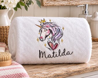 May include: A white rolled towel featuring an embroidered graphic of a unicorn with a gold crown and a pink heart. The unicorn has a flowing mane in shades of pink and purple. The name Matilda is embroidered in black script below the unicorn.