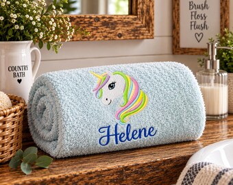 May include: A light blue rolled towel featuring an embroidered unicorn head with a multi-colored mane and horn. The name Helene is embroidered in dark blue script below the unicorn. The towel has a textured, plush surface.
