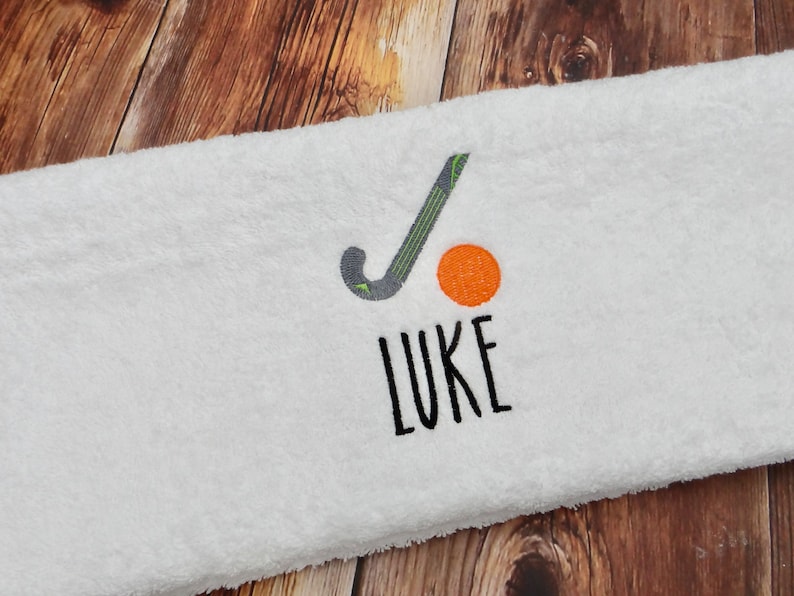 May include: White towel with embroidered hockey stick, orange ball, and the name "LUKE" in black.