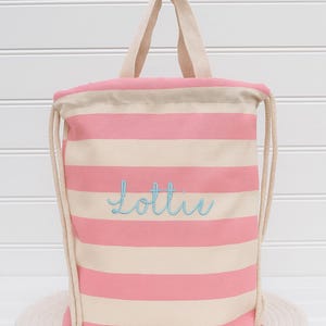Puede incluir: Una bolsa con cordón a rayas rosas y blancas con el nombre "Lottie" bordado en escritura azul claro. La bolsa tiene asas y cordones beige. Está sobre una estera tejida.