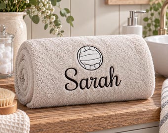 May include: A rolled beige bath towel featuring an embroidered black volleyball icon and the name Sarah in a cursive black script font. The towel has a soft, textured surface and is positioned horizontally on a wooden surface in a bathroom setting.