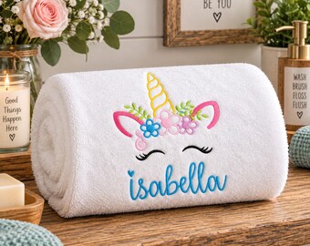 May include: A white rolled towel featuring an embroidered unicorn face with a yellow horn, pink ears, and a floral crown in blue and pink. Below the unicorn, the name Isabella is embroidered in blue script, accompanied by a small blue heart.