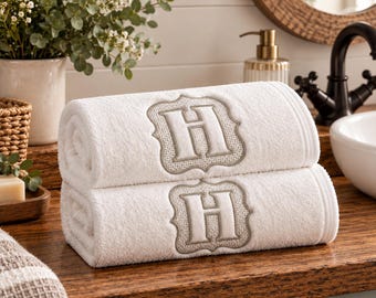 May include: Two stacked white cotton towels featuring a centered, embroidered beige letter H inside a decorative scalloped frame. The towels have a soft, textured pile and are neatly rolled, showcasing the monogram detail on the front surface.