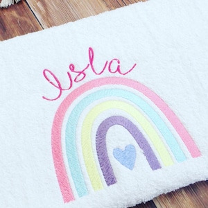 Op de afbeelding: Een witte handdoek met een geborduurde regenboog. De regenboog heeft roze, lichtblauwe, gele en paarse strepen. Een klein blauw hartje bevindt zich in het midden van de regenboog. De naam "Isla" is in roze boven de regenboog geborduurd.