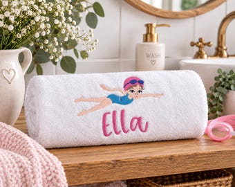 Könnte beinhalten: Ein weißes, gerolltes Handtuch mit einer Stickerei, die eine schwimmende Person in blauem Badeanzug und pinker Badekappe zeigt, darunter der Name Ella in pinker Schreibschrift. Das Handtuch hat eine weiche und flauschige Textur.