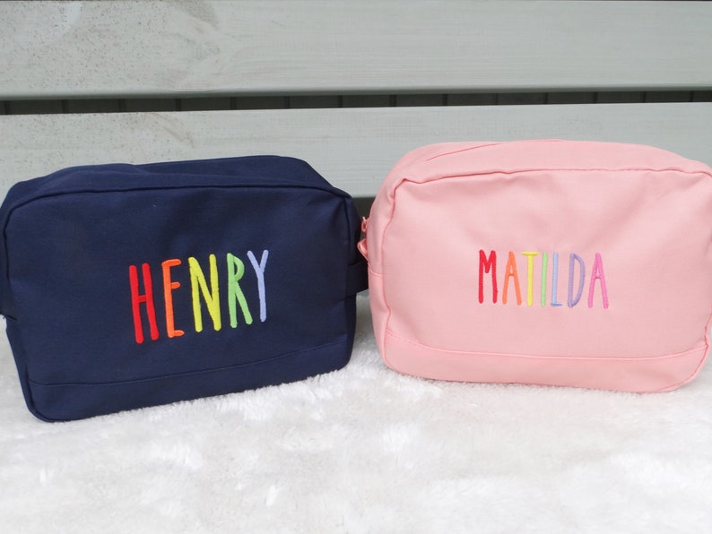 May include: Two personalised wash bags, one navy blue with the name "HENRY" embroidered in rainbow colours and one pink with the name "MATILDA" embroidered in rainbow colours.