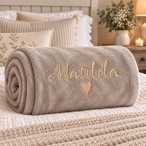 May include: A rolled, plush, taupe-coloured fleece blanket featuring the name Matilda embroidered in a cursive, light-gold script font, accompanied by a small, matching embroidered heart icon centred directly below the name on the fabric.