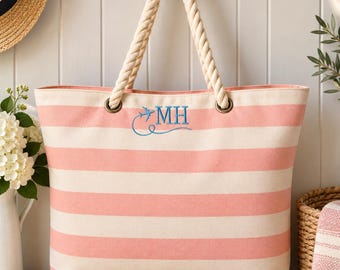 May include: A canvas tote bag featuring horizontal light pink and cream stripes. The bag has thick, twisted cream-coloured rope handles attached with metal grommets. Embroidered in blue on the front is a small aeroplane icon and the letters MH.