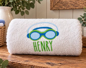 Könnte beinhalten: Ein weißes Frotteehandtuch mit einer Stickerei von blauen und grünen Schwimmbrillen mit einem hellblauen Band. Unter der Schwimmbrille ist der Name HENRY in leuchtend grünen Großbuchstaben in einer einfachen serifenlosen Schrift eingestickt.