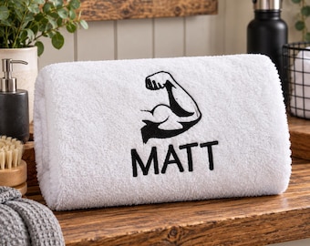 Può includere: Un asciugamano da bagno bianco con un ricamo nero raffigurante un braccio con bicipite flesso sopra il nome MATT in lettere maiuscole in grassetto. L'asciugamano è piegato in forma cilindrica, mostrando il tessuto morbido e strutturato.