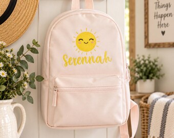 May include: A light pink backpack featuring an embroidered yellow sun with a smiling face and the name Serennah in yellow script lettering. The bag includes a top handle, adjustable shoulder straps, and a front zippered pocket with a matching pull.