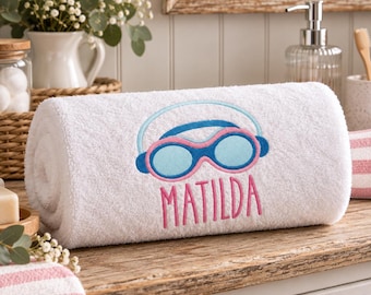 Könnte beinhalten: Ein weißes, gerolltes Handtuch mit einer Stickerei aus hellblauen und rosafarbenen Schwimmbrillen. Unter der Brille ist der Name MATILDA in rosa Großbuchstaben eingestickt. Das Handtuch hat eine weiche, strukturierte Oberfläche.