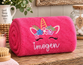 May include: A bright pink rolled towel featuring an embroidered unicorn face with a yellow horn, colorful floral accents, and the name Imogen stitched in white cursive script below the eyes. The towel has a soft, textured fabric surface.