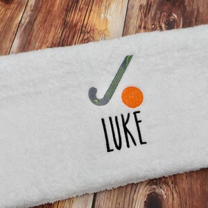 May include: White towel with a black and green hockey stick and an orange ball embroidered on it. The name "LUKE" is embroidered below the stick and ball.
