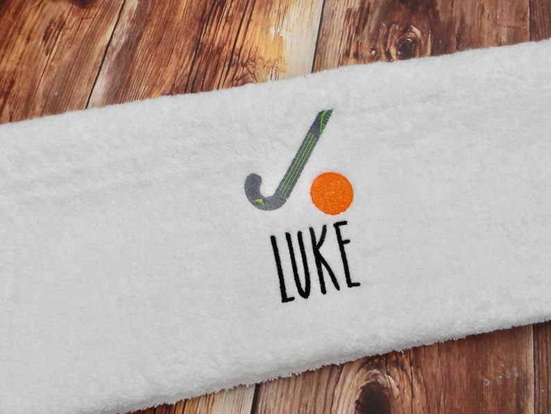 May include: White towel with a black and green hockey stick and an orange ball embroidered on it. The name "LUKE" is embroidered below the stick and ball.