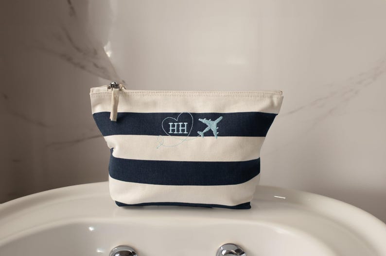 May include: A white and navy blue striped canvas zippered pouch with the initials "HH" embroidered in a heart shape and a blue airplane above it.