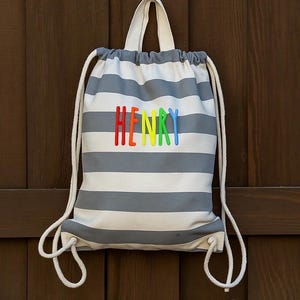May include: A grey and white striped drawstring bag with the name "HENRY" embroidered in rainbow colours. The bag has white rope straps and a white handle. The background is a dark brown wooden surface.