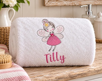 May include: A white rolled towel featuring an embroidered design of a fairy with a gold crown and a pink dress. Below the fairy, the name Tilly is embroidered in pink script. The towel has a textured surface and is made of soft cotton fabric.