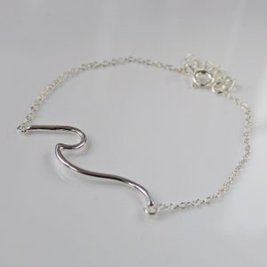 Pode incluir: Um bracelete de prata com forma de onda e uma corrente delicada. A onda é um design simples e abstrato.