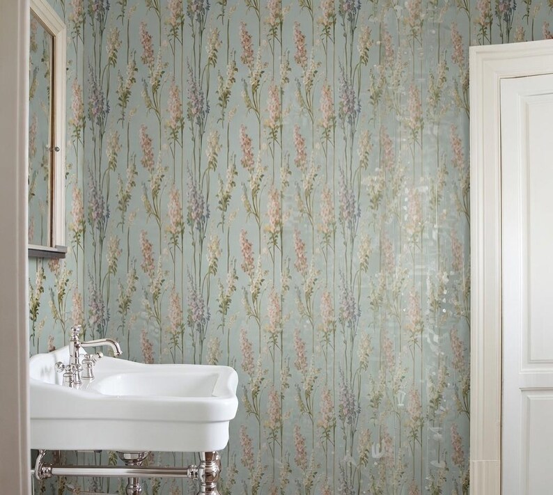 May include: A bathroom with floral wallpaper in shades of pink, purple, and cream on a light blue background. A white porcelain sink with chrome fixtures is in the foreground. A white door and mirror are also visible.